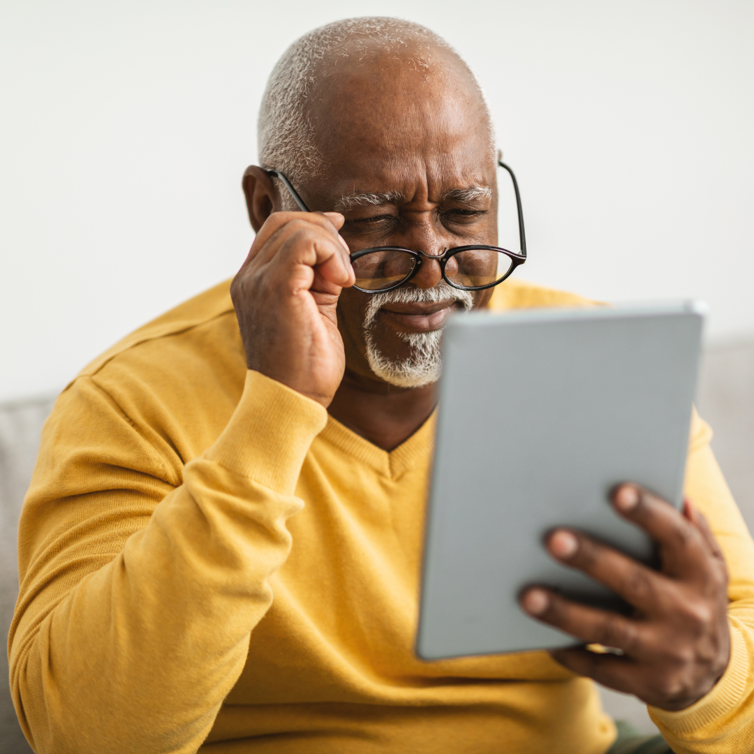 Man wearing a yellow top and glasses. Holding a tablet with his glasses pushed down on to his nose struggling to read his tablet.